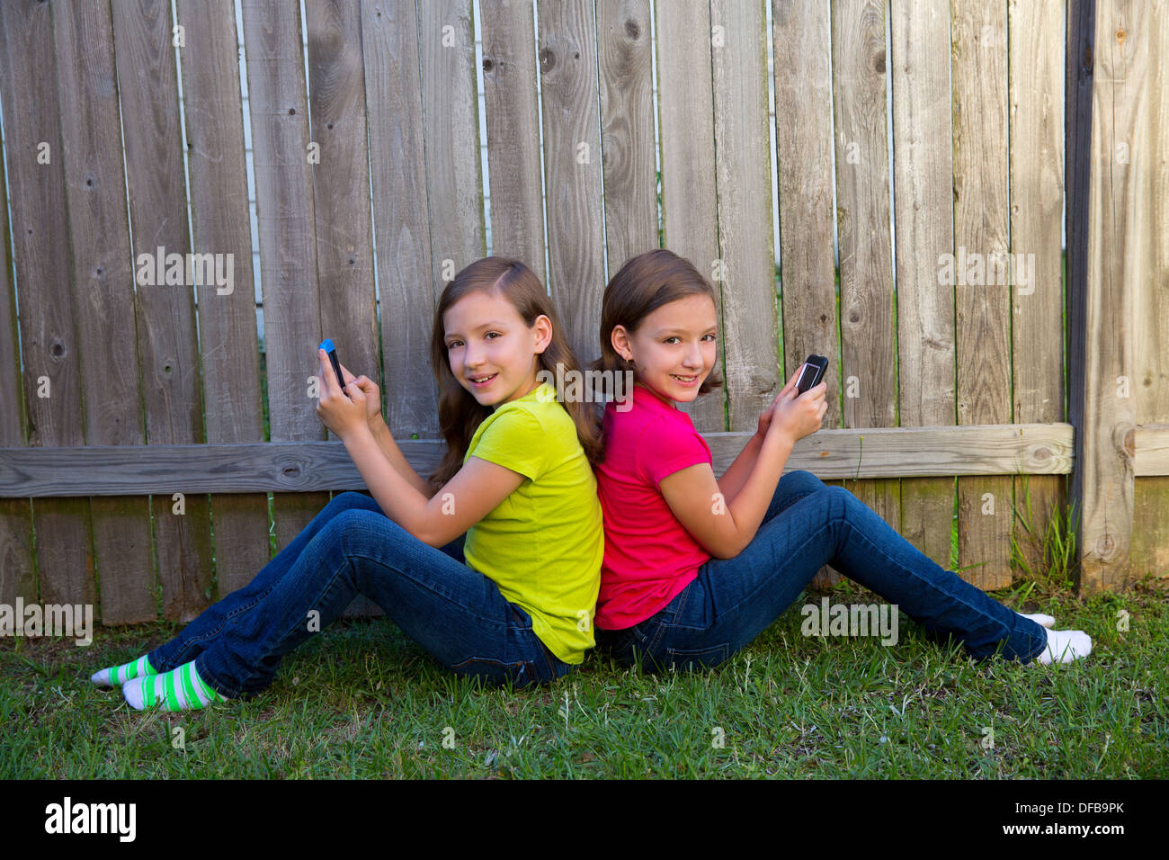 Twin sister girls playing with smartphone sitting on backyard lawn fence leaning on her back Stock Photo