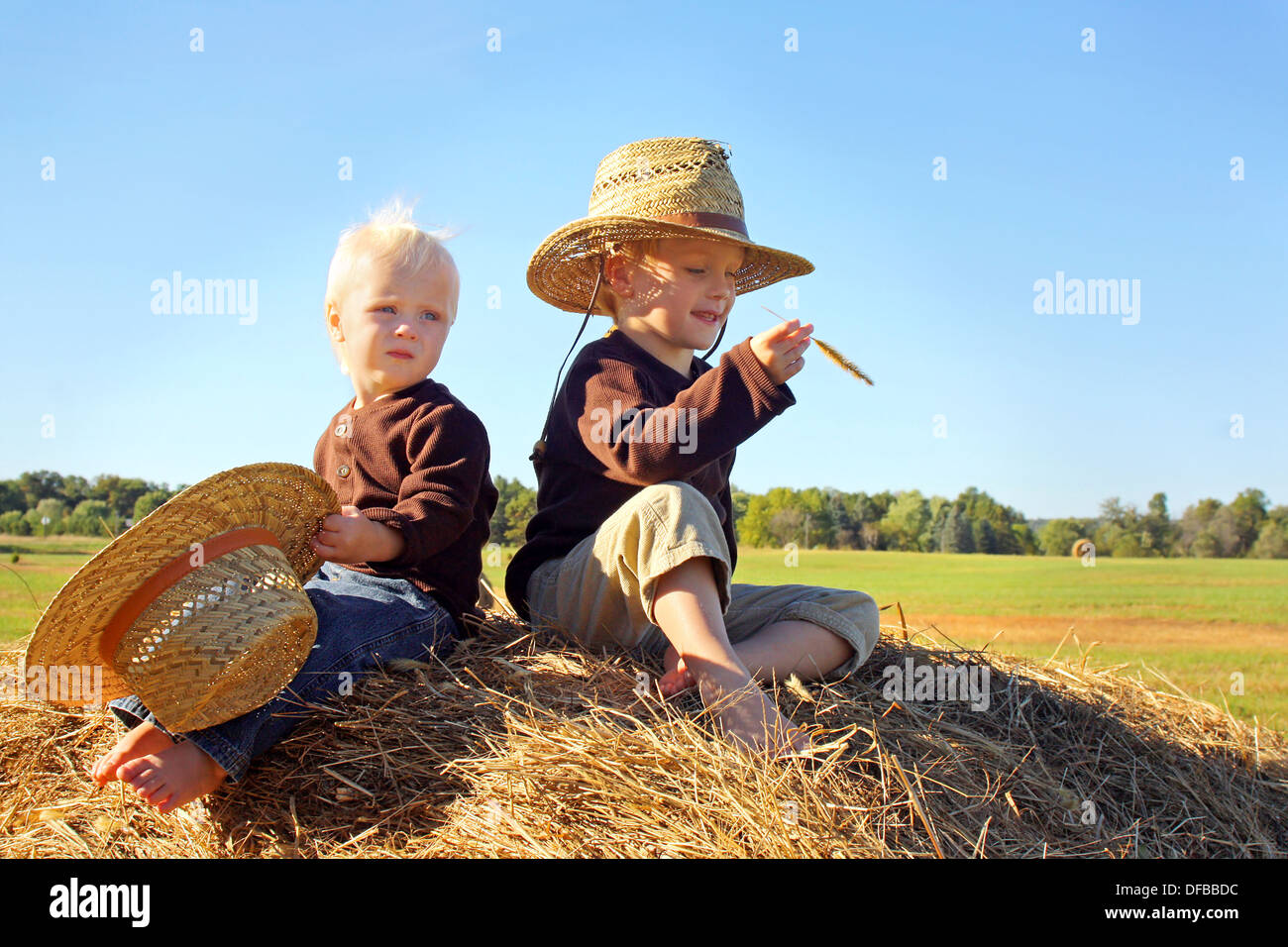 Two young kids, a little boy and his baby brother, are sitting on top of a hay bale wearing straw hats on a sunny autumn day. Stock Photo