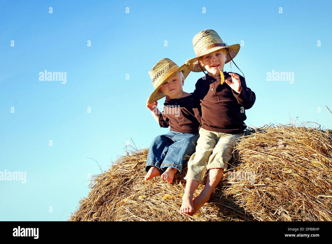 Two children, a young boy and his baby brother are sitting outside on hay bales, wearing straw hats on a sunny Autumn day. Stock Photo