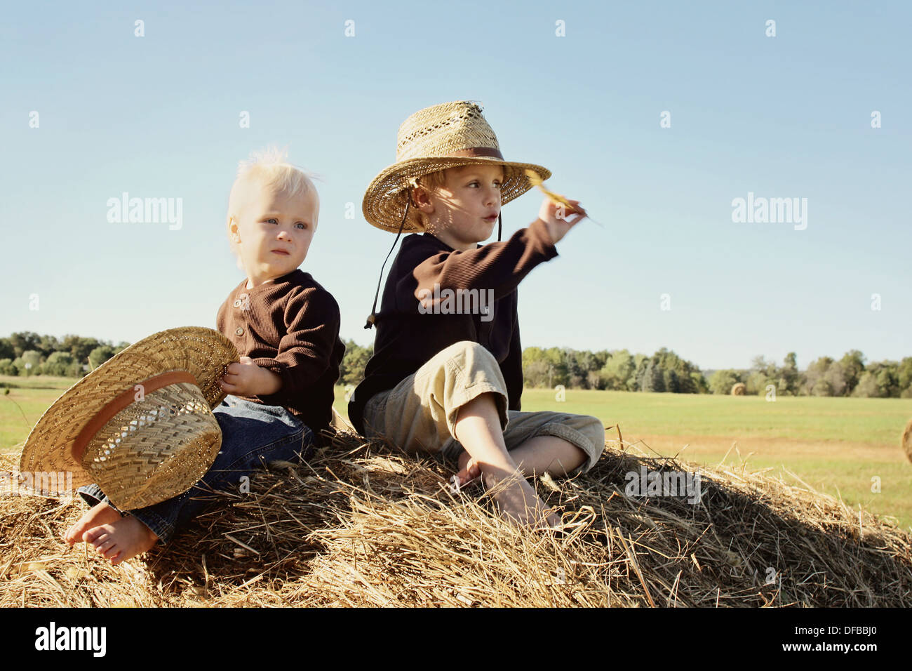 Two happy little boys, a 4 year old and baby brother, are sitting on top of a large hay bale, wearing straw hats on autumn day Stock Photo