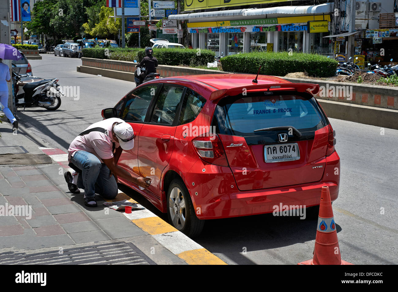 Man undertaking roadside repairs and respraying paintwork on a red Honda Jazz using a spray can. Thailand S. E. Asia Stock Photo