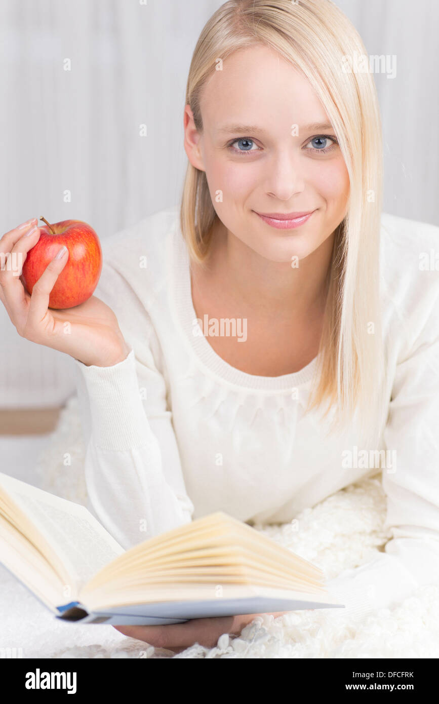 Portrait of young woman lying on couch and reading book with red apple ...