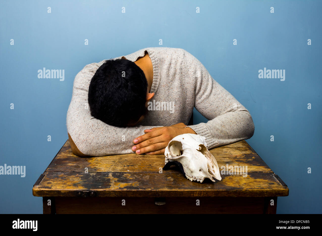 Sad young man is sat at a desk with a goat's skull Stock Photo
