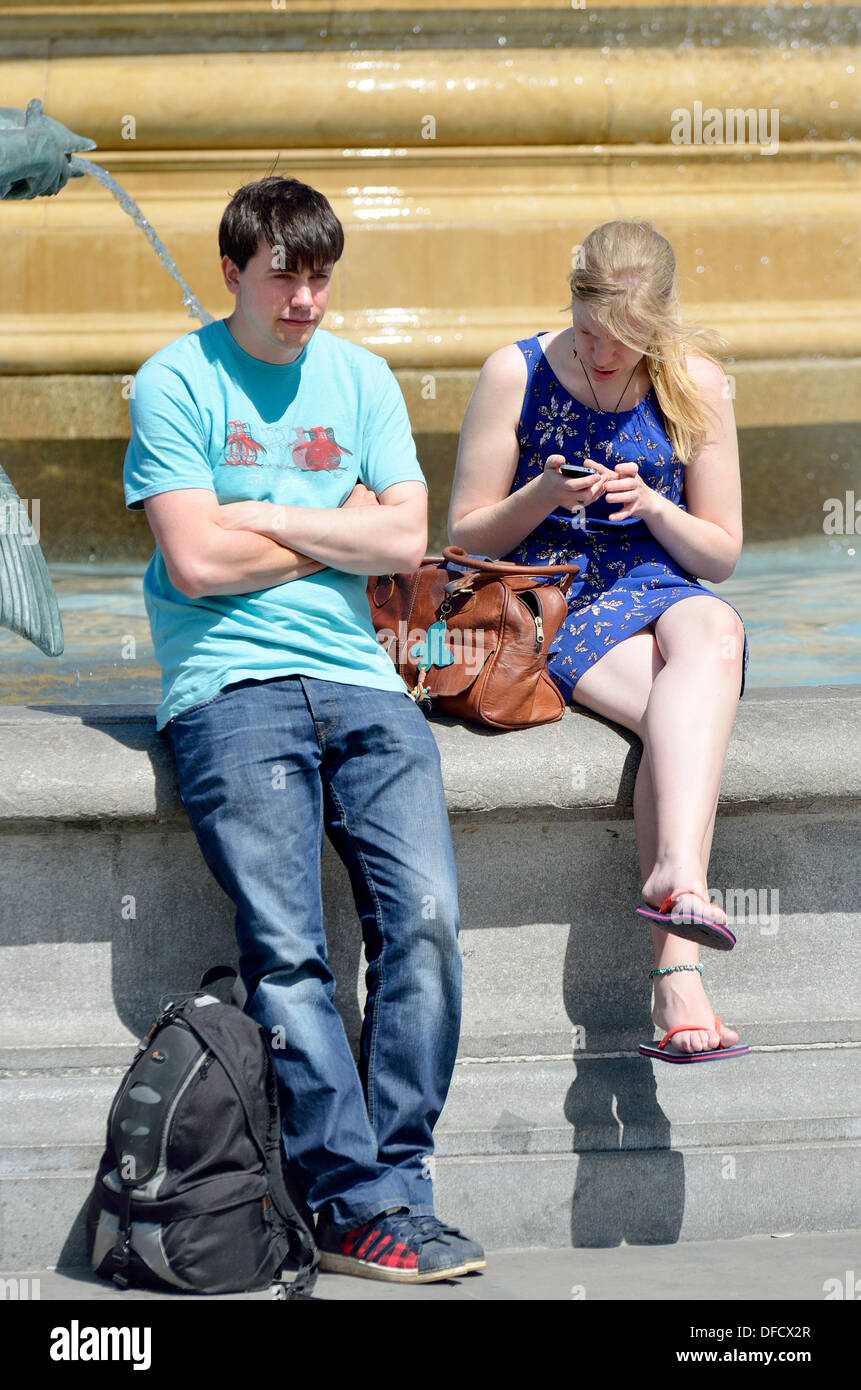 London, England, UK. Young couple (? no MR) by a fountain, girl on her phone, boy waiting Stock Photo