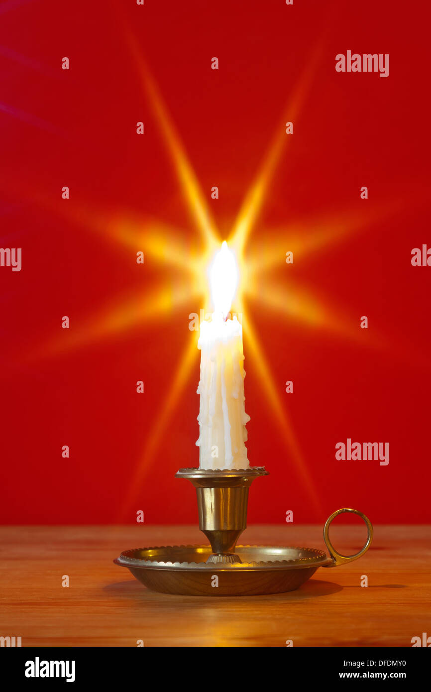 A dripping wax candle in a traditional brass holder known as a chamberstick, burning against a red background. Stock Photo
