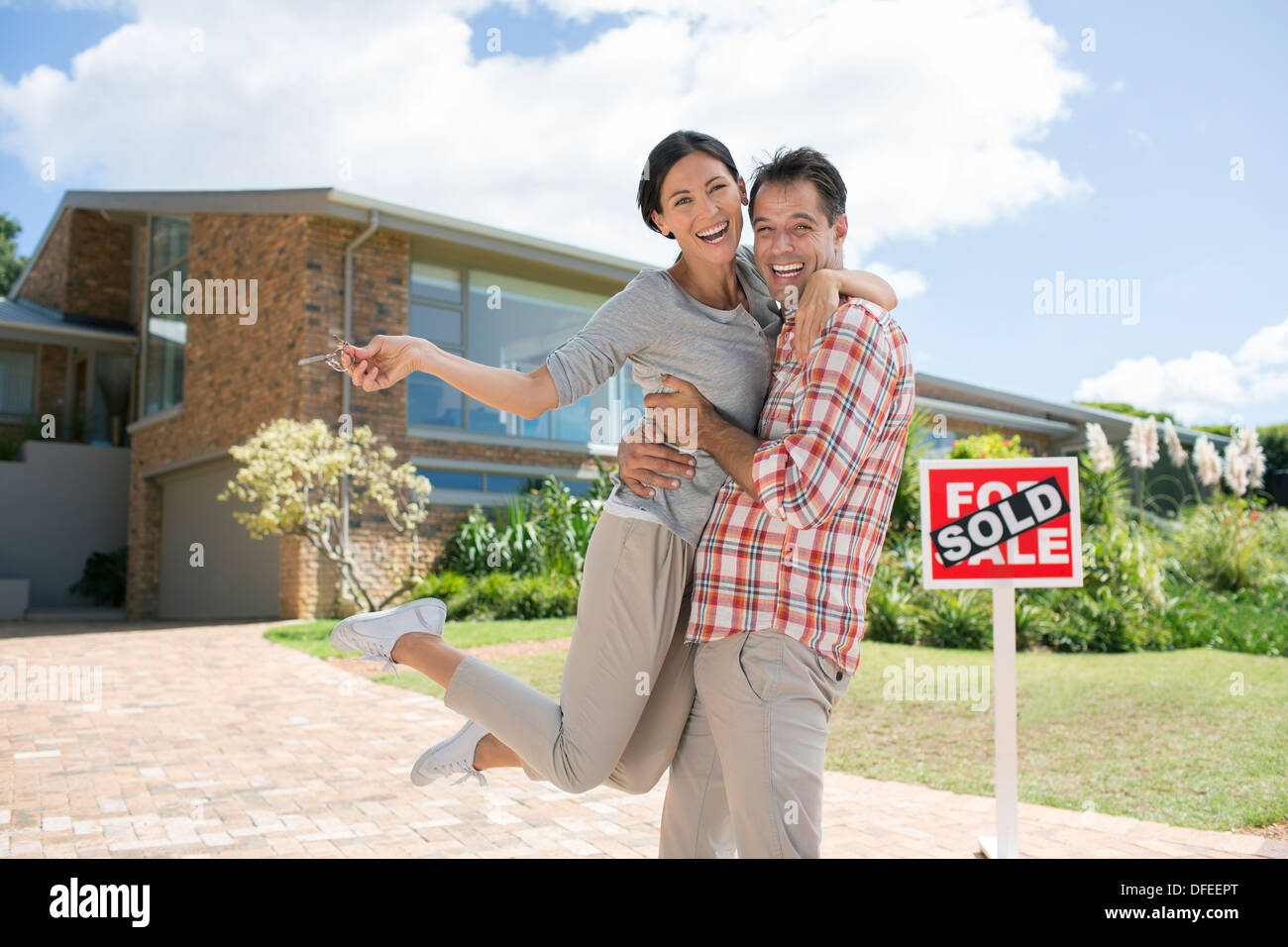 Portrait of enthusiastic couple hugging outside house with For Sale sign Stock Photo