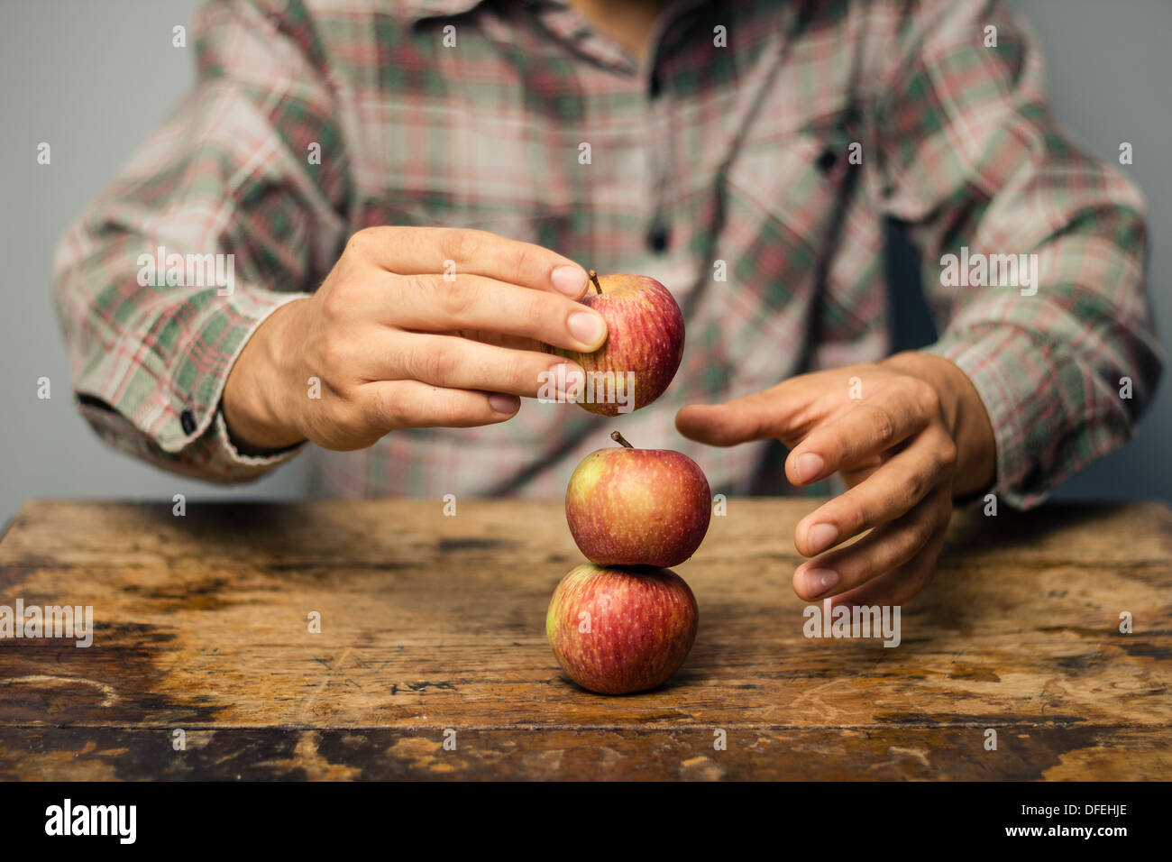 Young man is sitting at an old wooden table and stacking three apples on top of each other, to symbolize a balanced diet Stock Photo