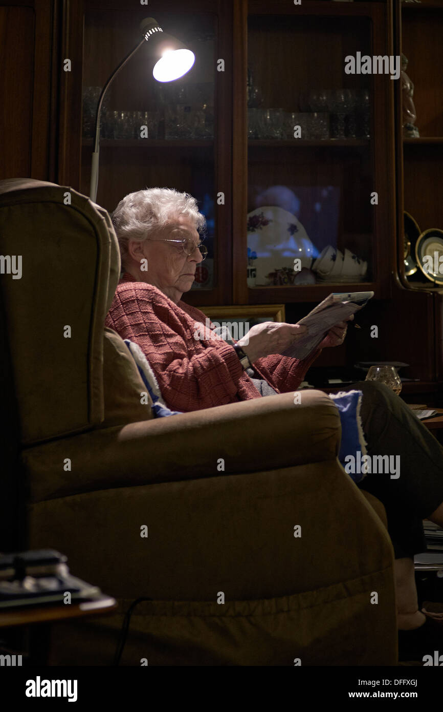 Elderly woman relaxing at home and reading a newspaper under a single spot light. Stock Photo