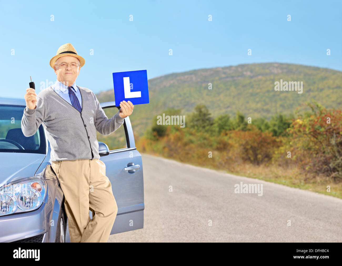 Happy senior man posing on his car, holding a L sign and car key after having his driver's license on an open road Stock Photo