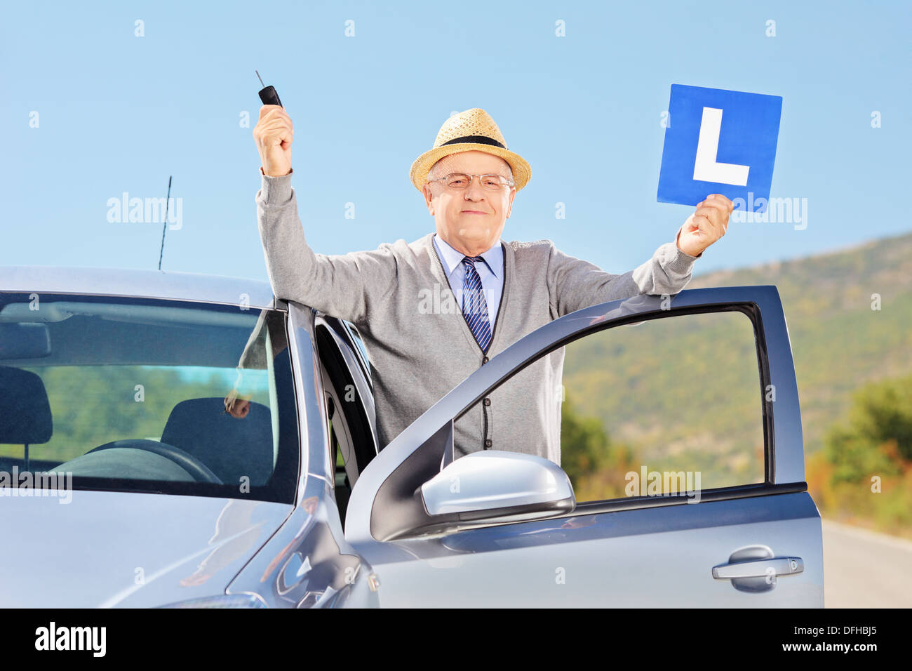 Smiling senior man posing next to his car holding a L sign and car key after having his driver's license Stock Photo