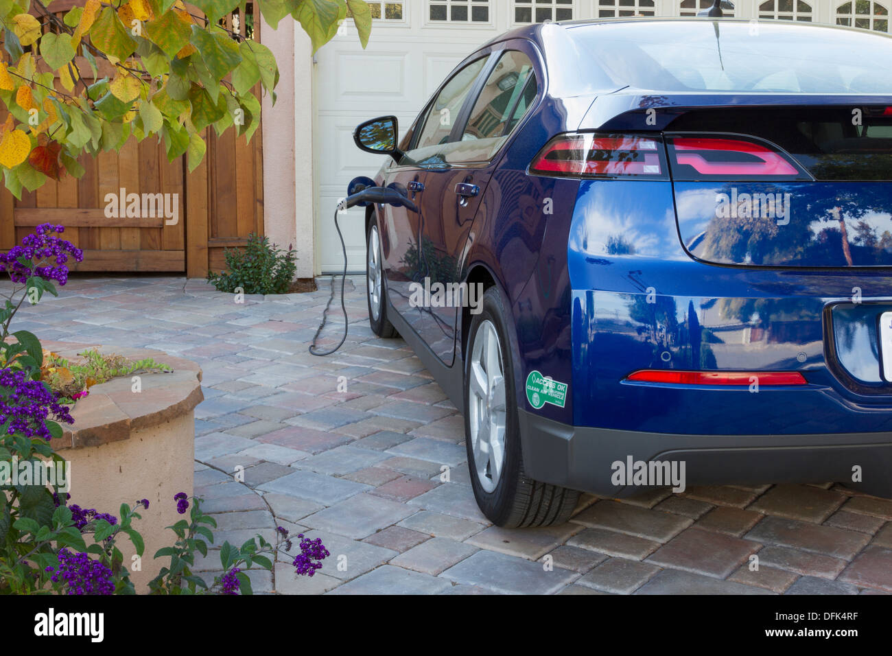 Plug-in electric car with carpool sticker parked in driveway, with connector plugged in and charging at home Stock Photo