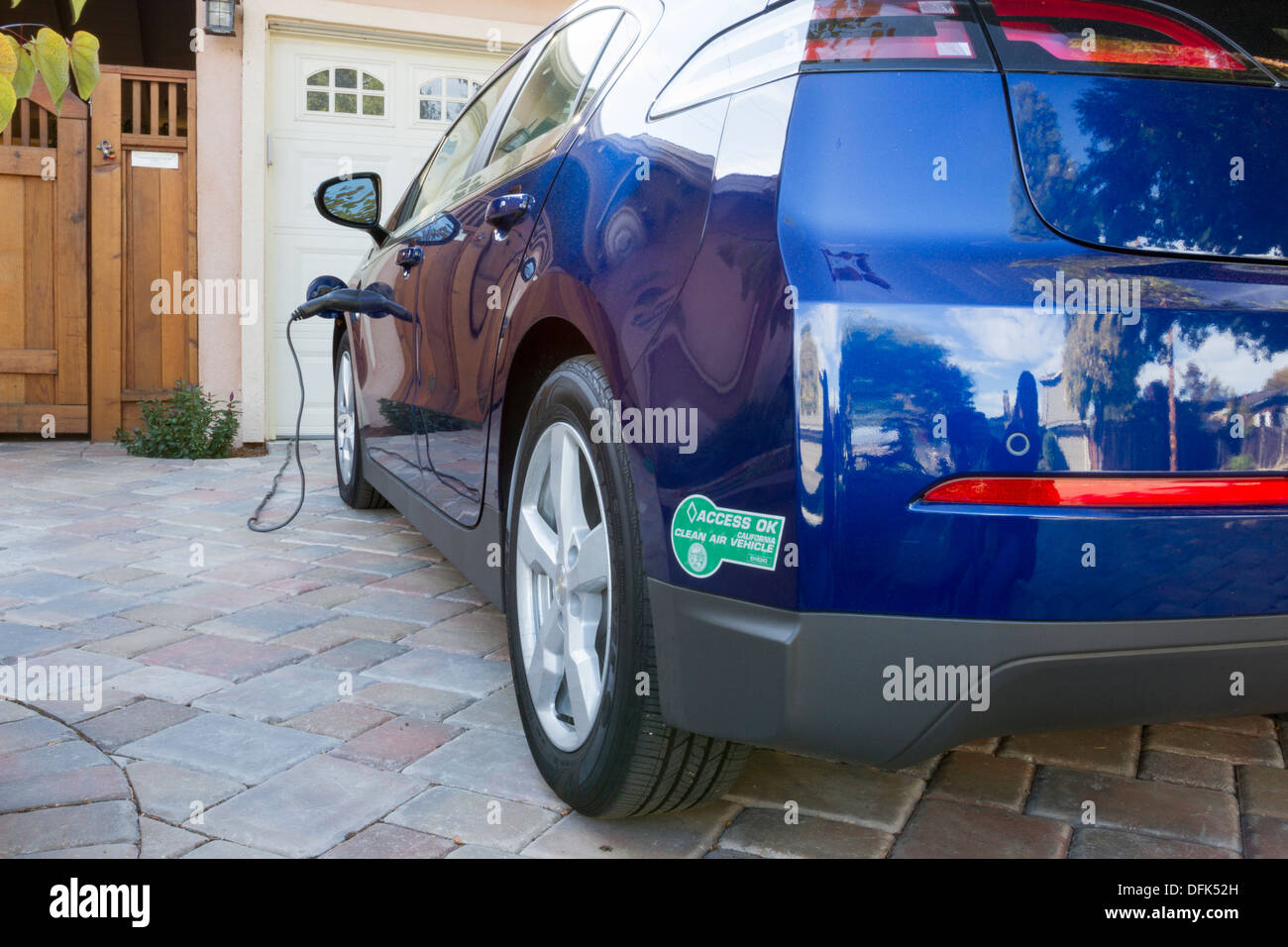 Plug-in electric car with carpool sticker parked in driveway, with connector plugged in and charging at home Stock Photo
