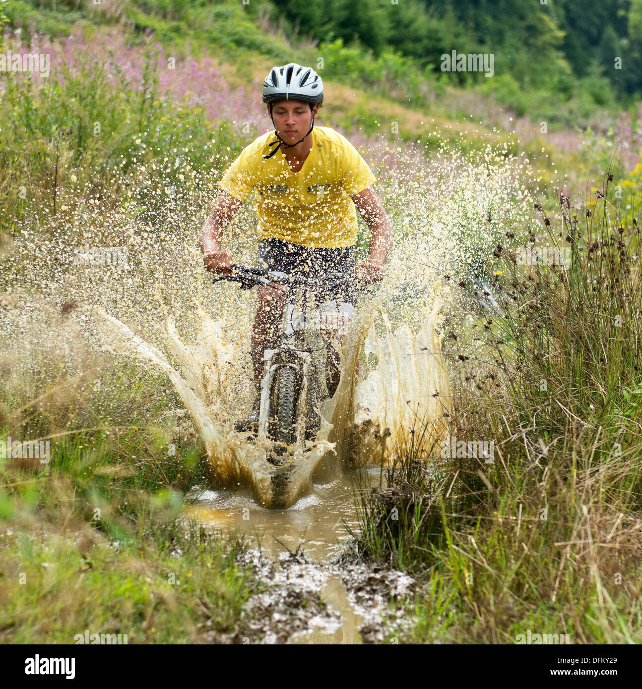 A young man on a mountain bike riding through a muddy puddle in grassy landscape Stock Photo