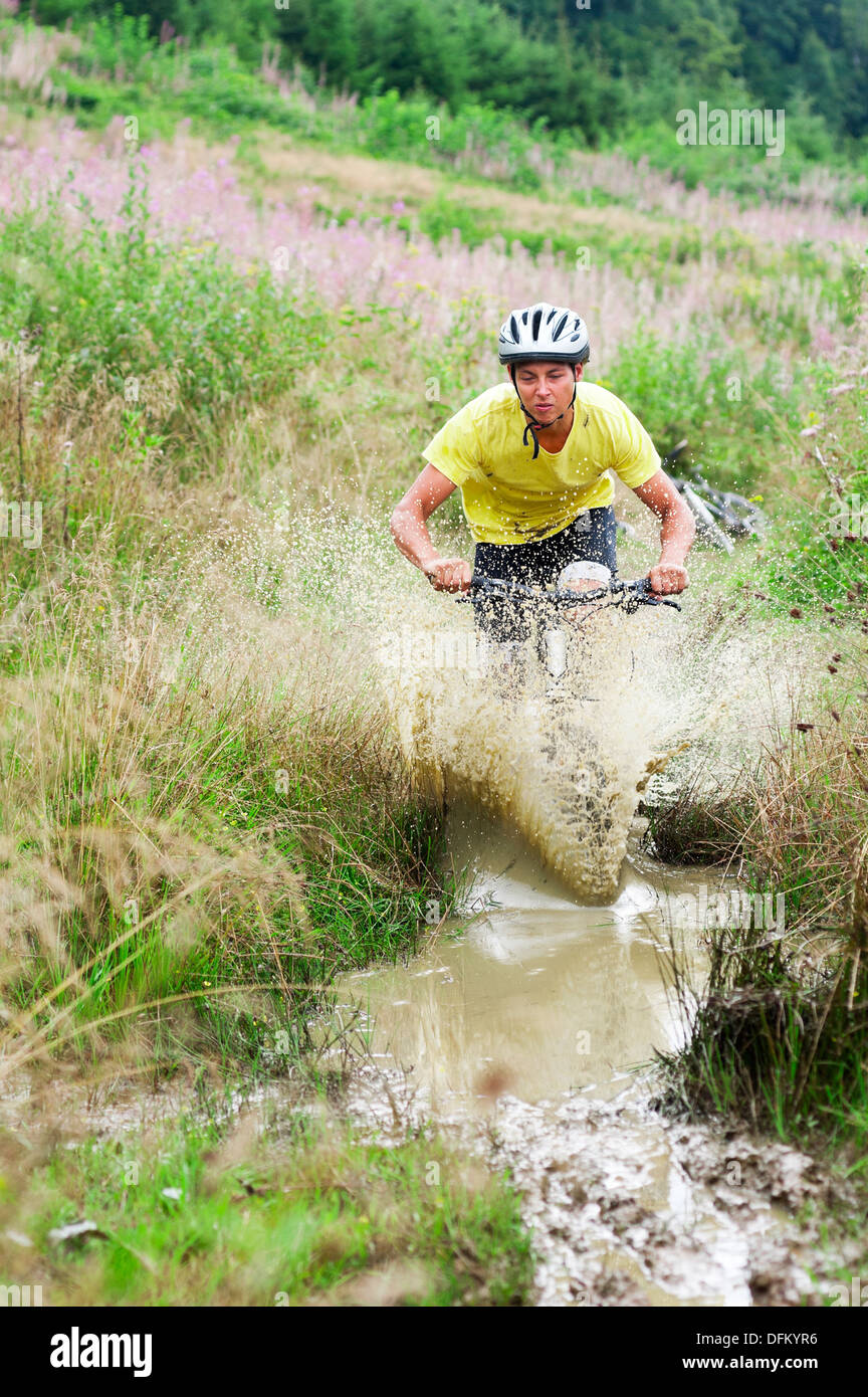 A young man on a mountain bike riding through a muddy puddle in grassy landscape Stock Photo