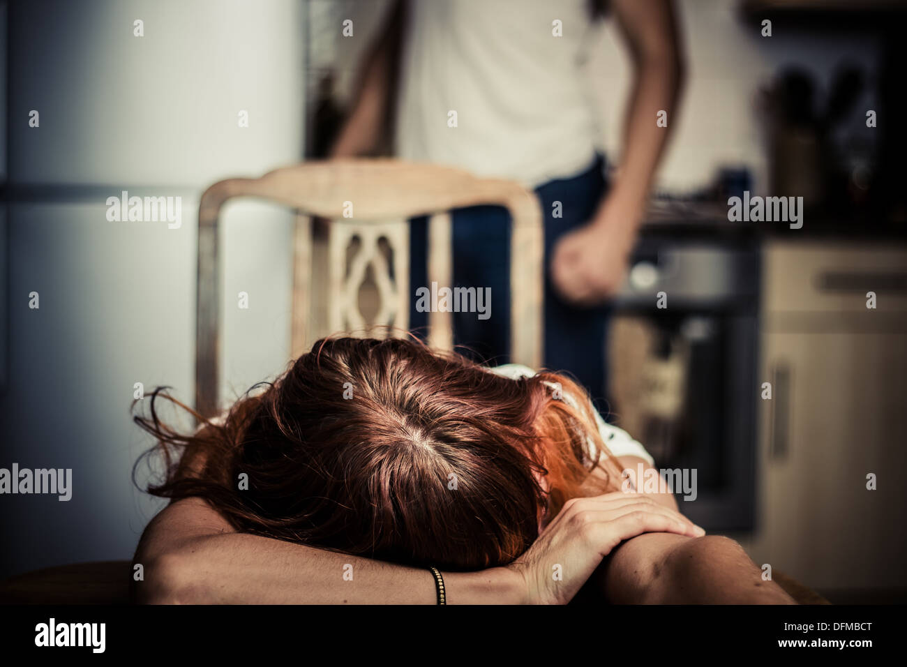 Young woman at table with abusive partner in background Stock Photo