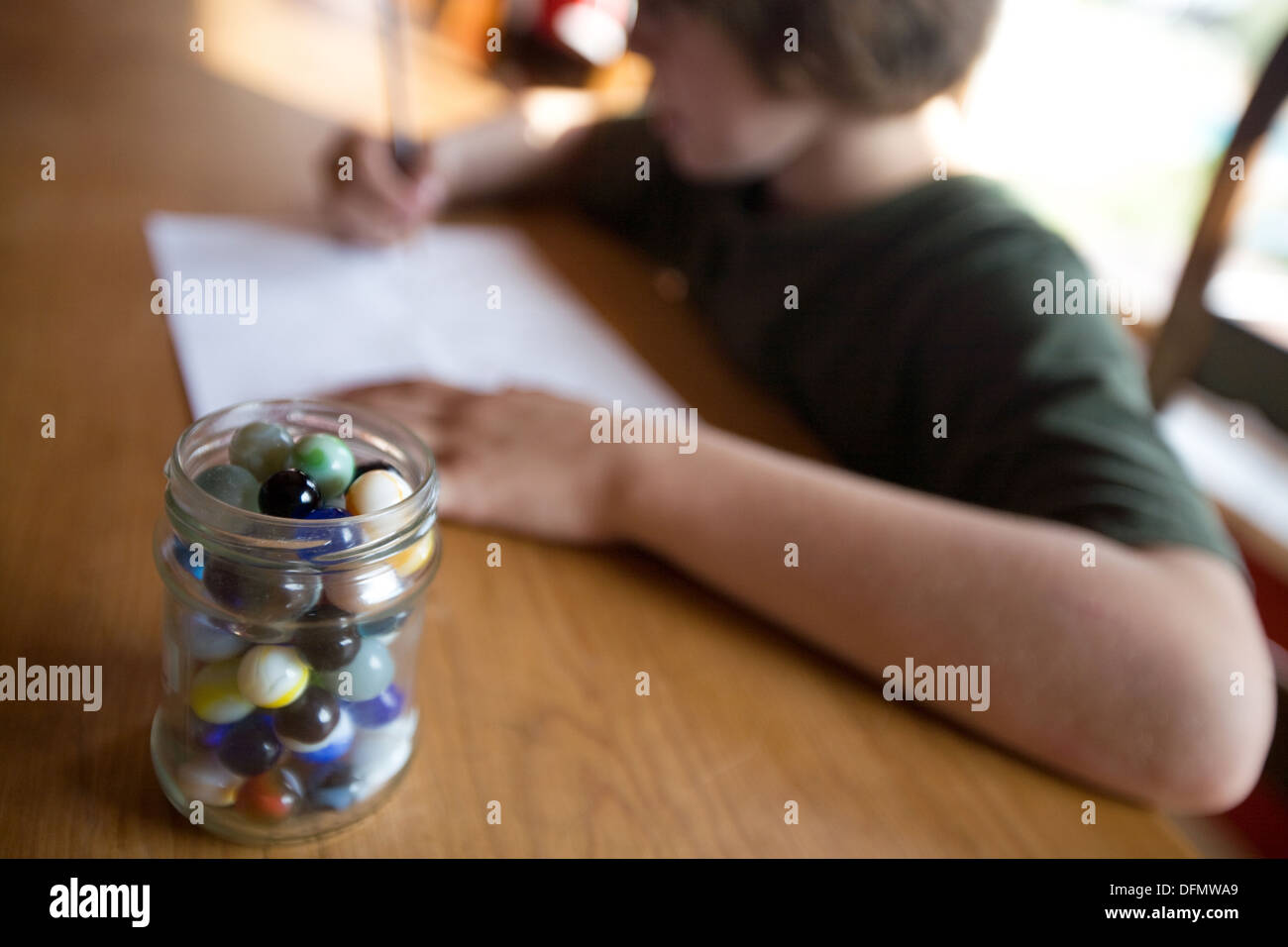 Boy concentrating on drawing at a table, a jar of marbles in foreground. Stock Photo
