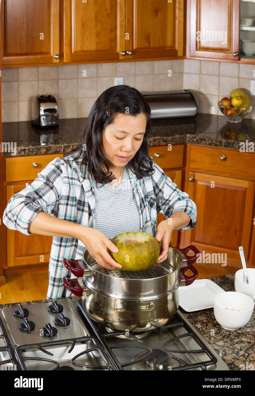 Vertical photo of mature woman lifting winter melon, from steaming pot, with both hands to place on white dinner plate Stock Photo