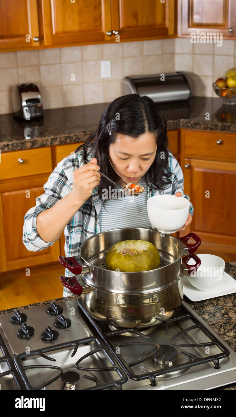 Vertical photo of mature woman tasting winter melon dinner, from steaming pot, with large spoon in one hand and a white bowl i Stock Photo