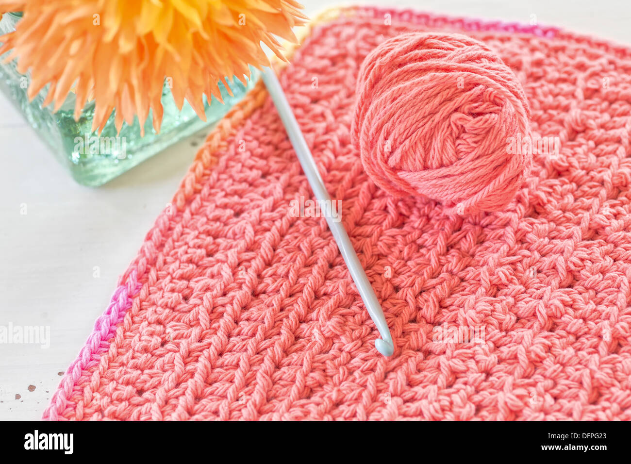 A crocheted dishcloth with crotchet hook and ball of yarn sitting alongside a vase of flowers. Stock Photo