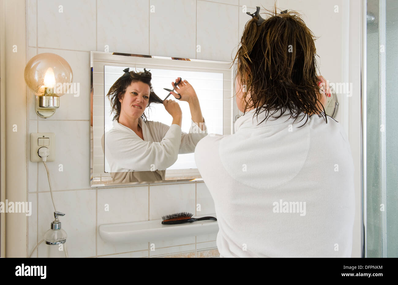 A woman is looking in the mirror of the bathroom and having a 'bad hair day', and wants to cut her hair Stock Photo