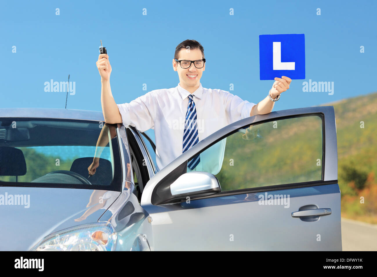 Happy young man posing near his car, holding a L sign and key after having his driver's licence on a road Stock Photo