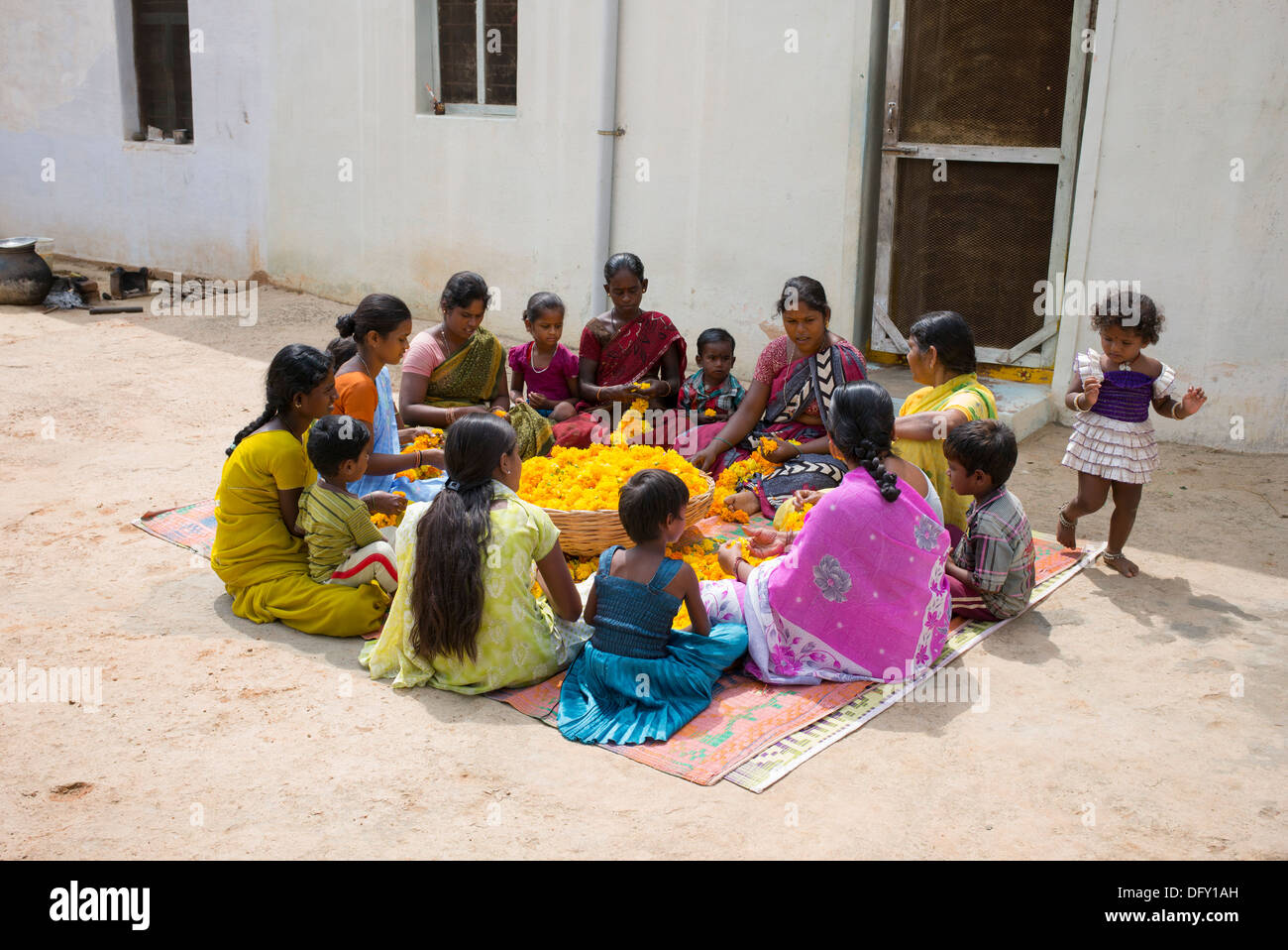 Rural Indian village women and children sitting round a basket of marigold flowers making flower garlands. Andhra Pradesh, India Stock Photo