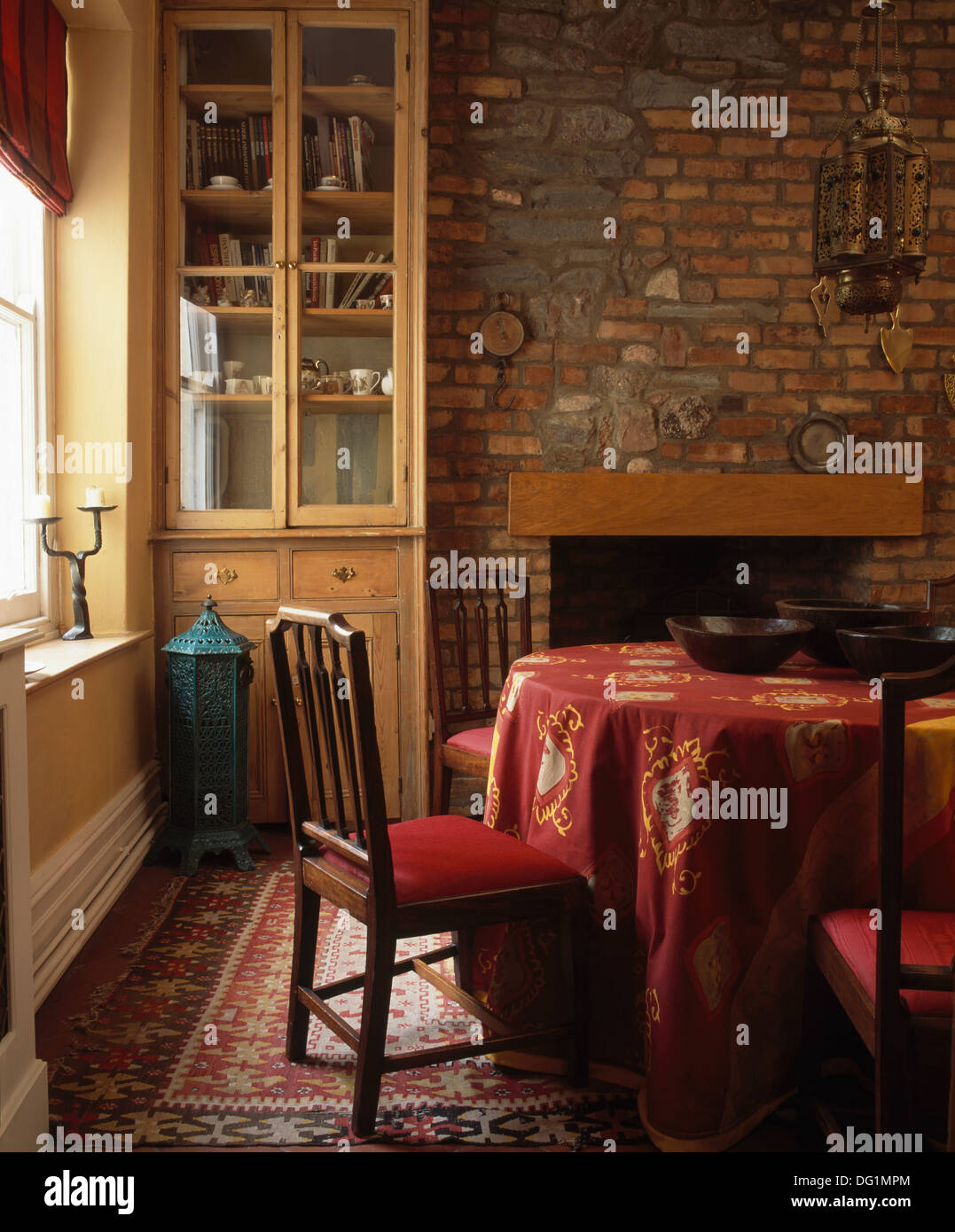 Narrow dresser and table with patterned red cloth in traditional dining room with exposed brickwork above the fireplace Stock Photo