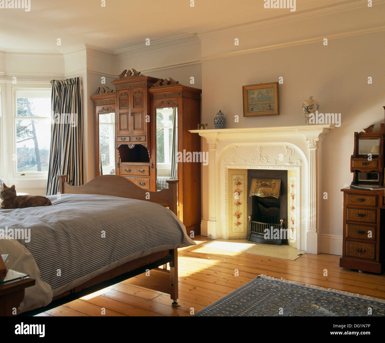 Large Edwardian wardrobe and white painted fireplace in pale blue bedroom with polished floorboards and bed with striped duvet Stock Photo