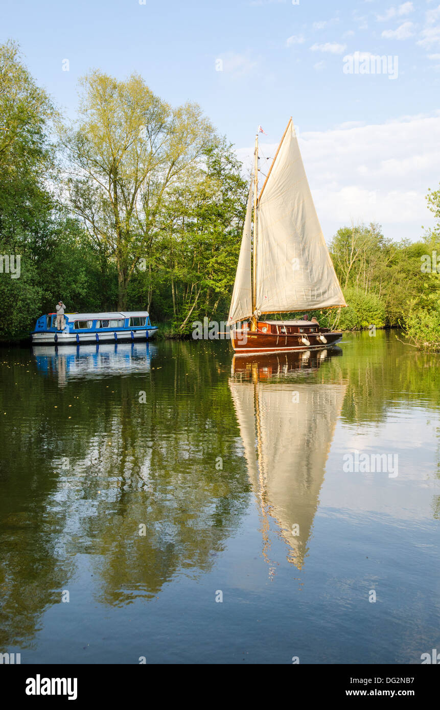 yacht sailing up River Bure, Norfolk Broads, England, past holiday launch houseboat moored under trees. Stock Photo