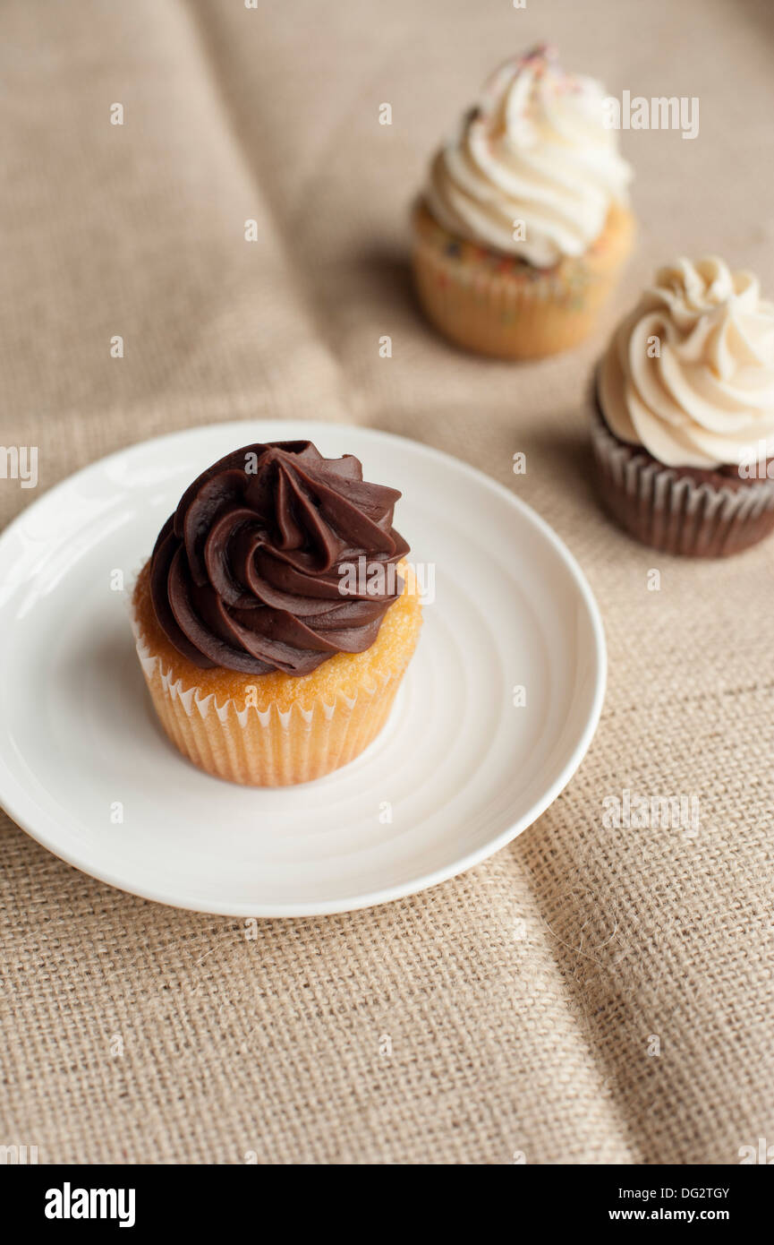 Cupcake with Chocolate Icing on White Plate and Two Cupcakes with Vanilla Icing in Background Stock Photo