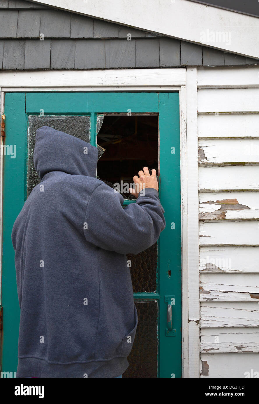 A suspicious person, man wearing a hooded jacket peering through a broken window pane in a door of a garden shed. Stock Photo