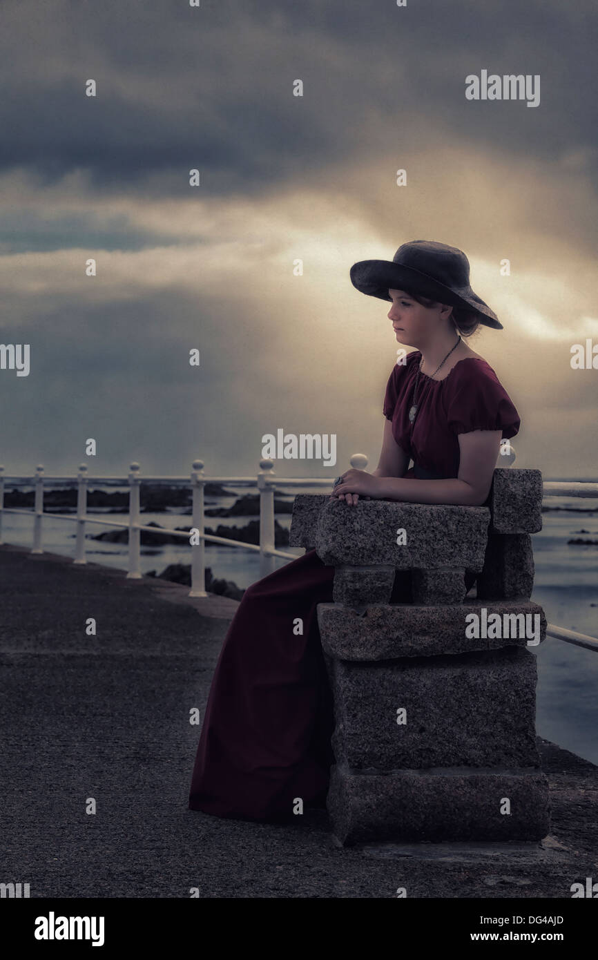 a sad girl with a black sunhat sitting on a stone bench at the sea Stock Photo