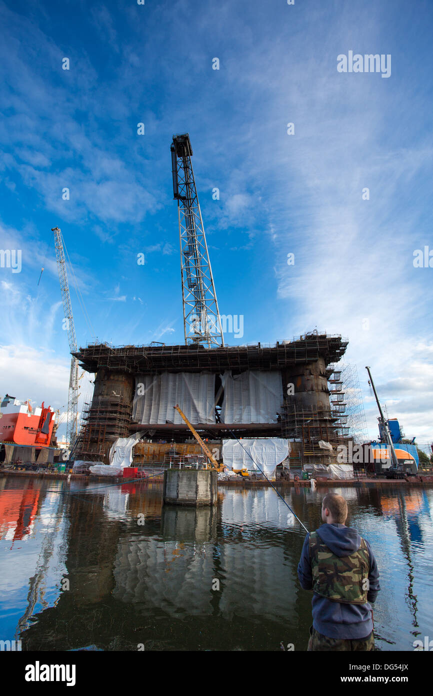 Fisher man fishing in front of a docking oil rig at the Gdansk Shipyard under construction with a clear blue sky. Stock Photo