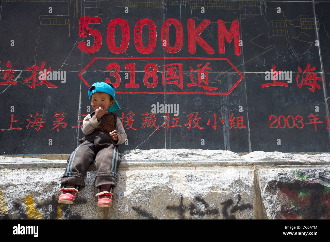 Tibetan Kid in front of a road sign.The Signs tell that we are exactly 5000 Km from Shanghai, on the Friendship Highway In Tibet Stock Photo