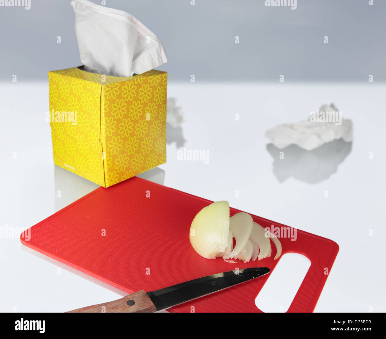 we see slices of an onion on a cutting board and a knife, a crumpled tissue, and  a box of tissues nearby Stock Photo