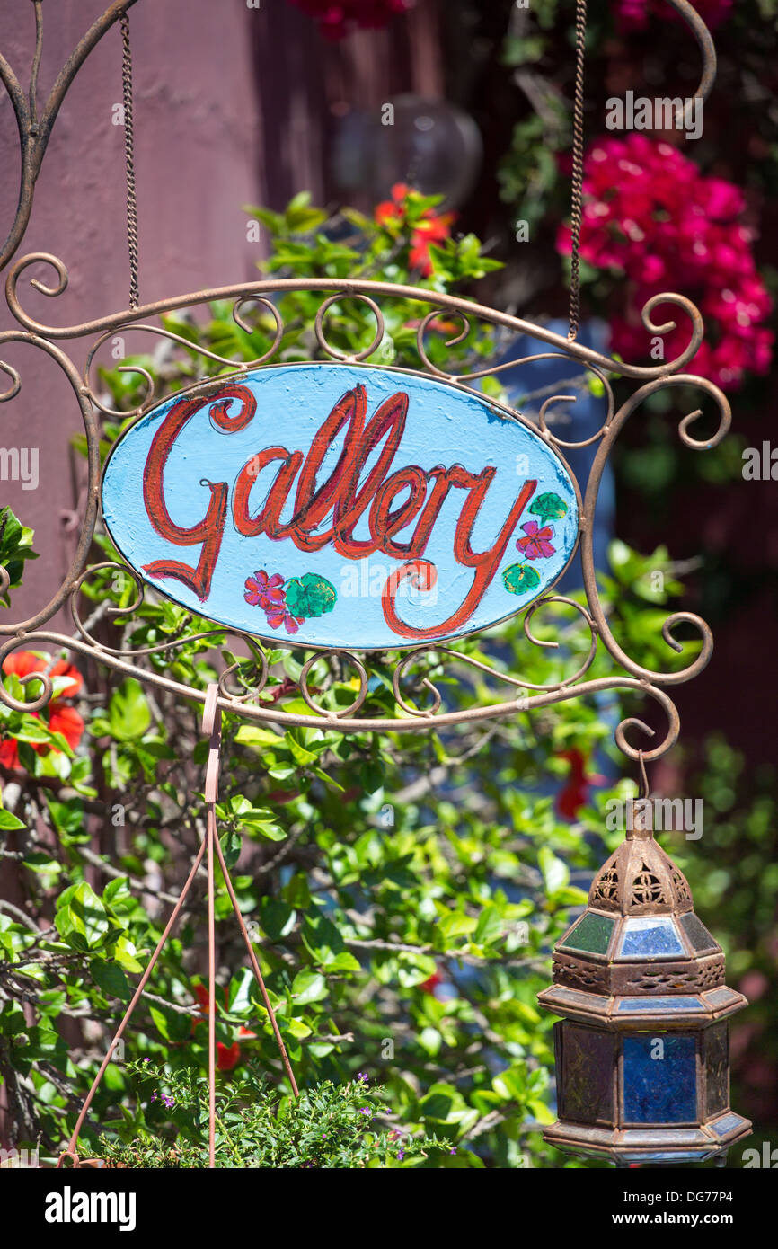 Blue sign board displaying the word gallery in red and surrounded by a green garden in Santorini Stock Photo