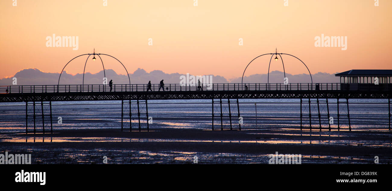 Silhouetted figures of people walking at sunset down Southport Pier with the ornate lights lit and tram shelter. Stock Photo