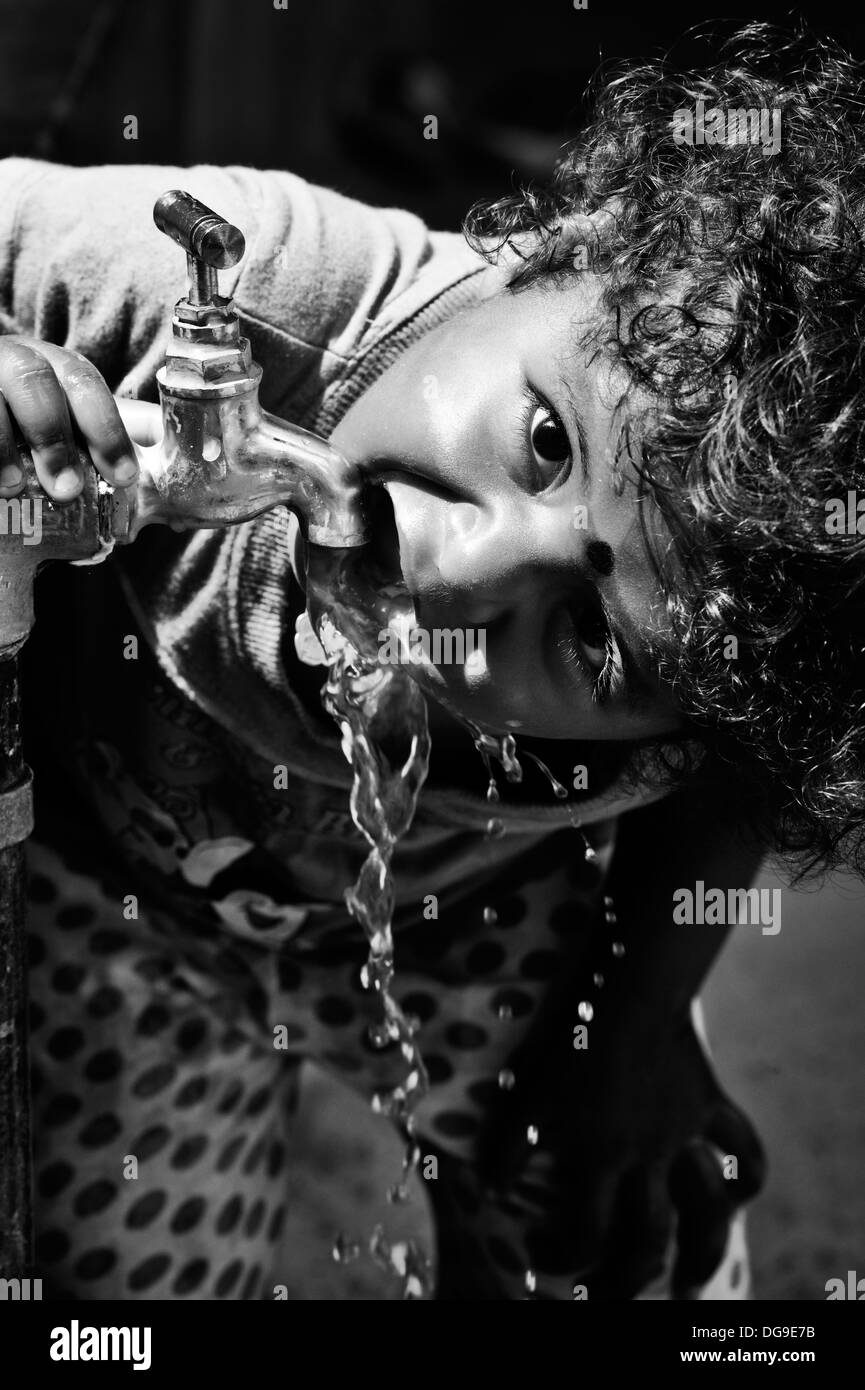 Young Indian girl / infant drinking water from a tap in a rural Indian village. Andhra Pradesh, India. Black and White Stock Photo
