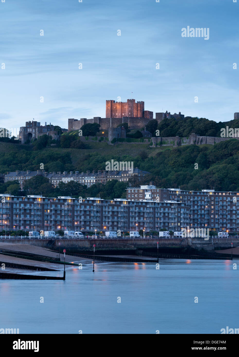 Dover Castle illuminated at dusk, above a row of buildings and the coastline below. Stock Photo