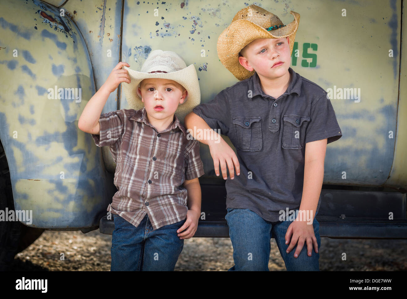 Two Young Boys Wearing Cowboy Hats Leaning Against an Antique Truck in a Rustic Country Setting. Stock Photo