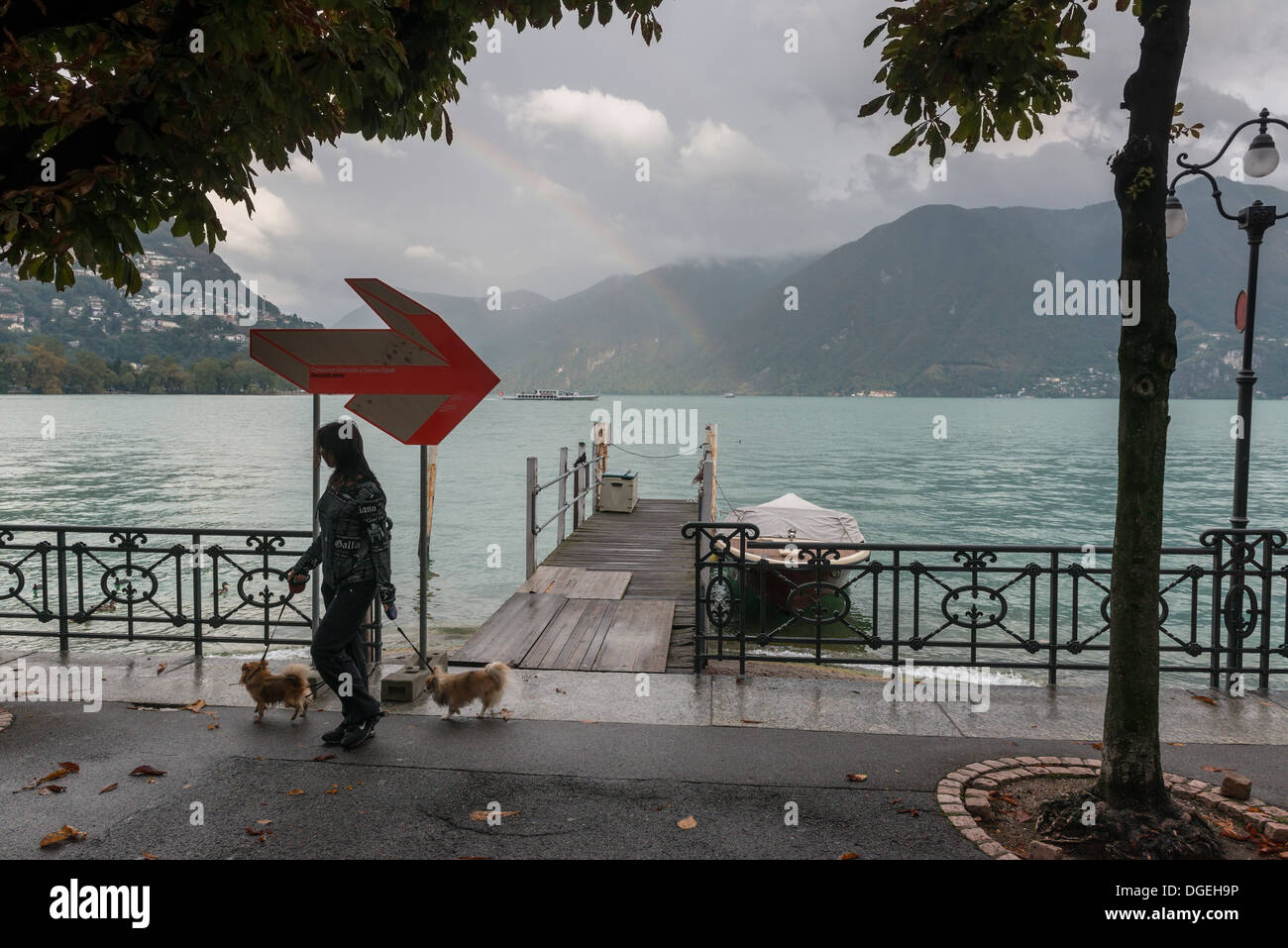 A female young adult walking her two dogs past a rainbow over Lake Lugano, Ticino, Switzerland Stock Photo