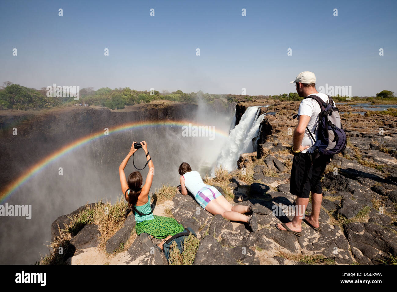 Three tourists looking at the Victoria Falls and rainbow from Livingstone Island, Zambia africa Stock Photo