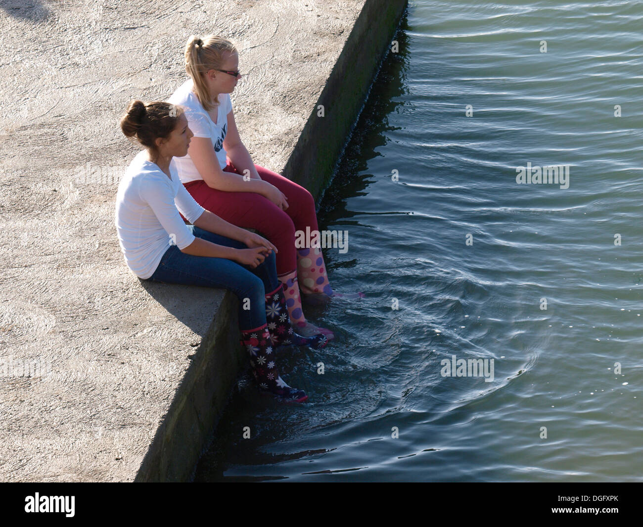 Two teenage girls sat on the edge of seapool dangling feet in the water, Bude, Cornwall, UK Stock Photo