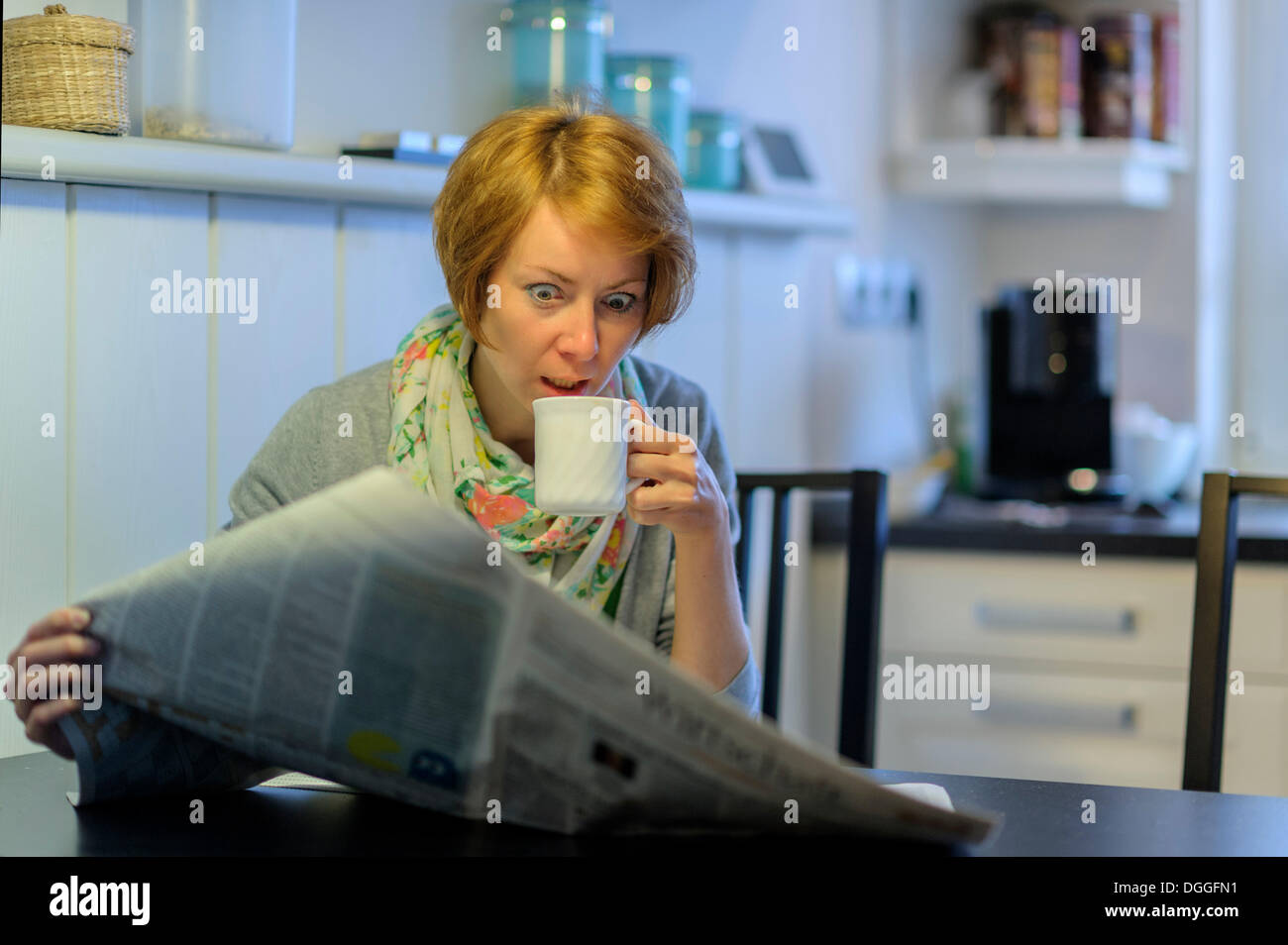 Woman sitting at the kitchen table reading the newspaper, with an astonished look, Germany Stock Photo