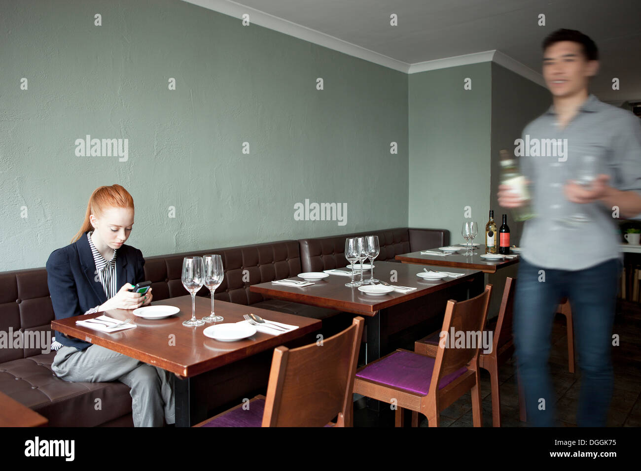 Young woman using cell phone in restaurant, waiter walking past Stock Photo