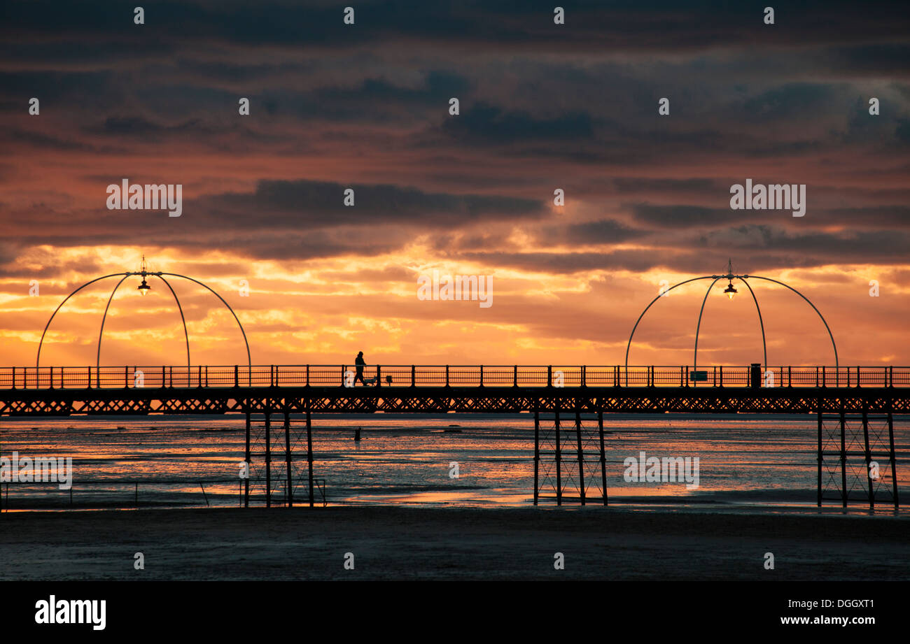 Silhouette figure of man walking his dog at sunset down Southport Pier with the ornate lights lit and tram shelter. Stock Photo