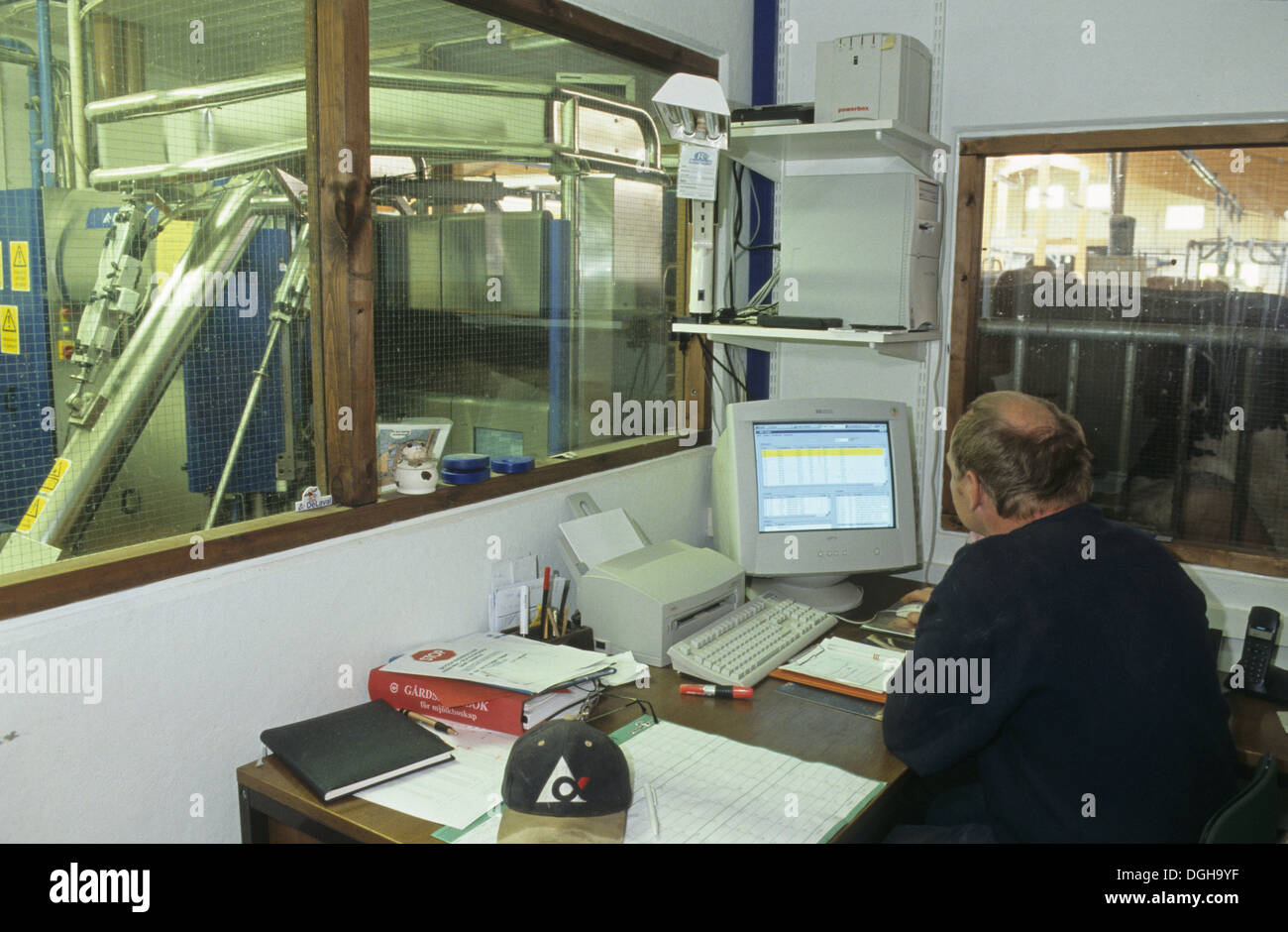 Dairy farming, farmer sitting at desk in office, using computer, milking machine beyond internal window, Sweden Stock Photo