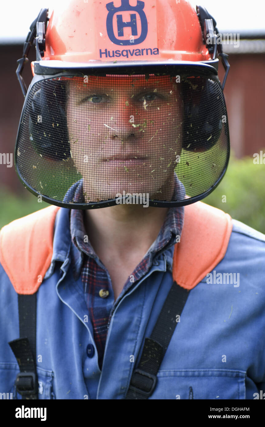 Man wearing protective helmet with visor and ear defenders, using strimmer on farm, Sweden Stock Photo