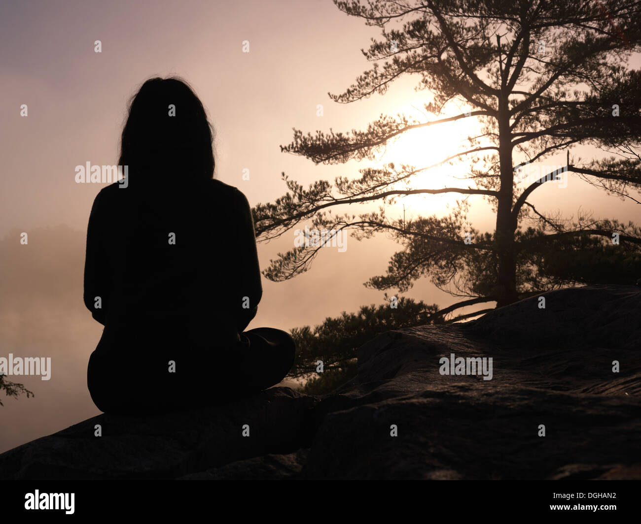 Silhouette of a woman sitting on rocks and facing sunrise sun, tranquil nature scenery, relaxation and meditation concept Stock Photo