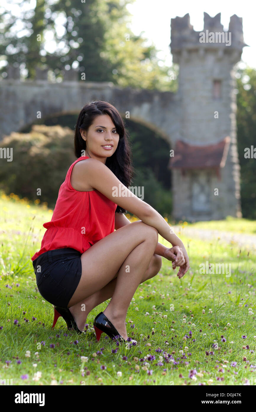 Young woman in a red top, black hot pants and high heels posing on a meadow in front of a castle tower Stock Photo